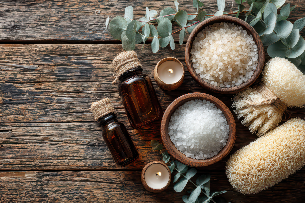 Essential oil bottles, bowls of salt and sugar, and candles on a wooden surface with eucalyptus leaves.
