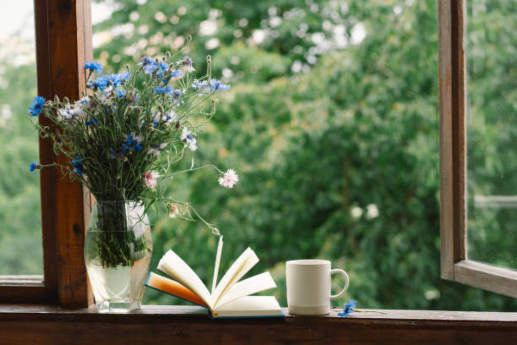 Open book, cup, and vase of flowers on a windowsill with greenery outside.