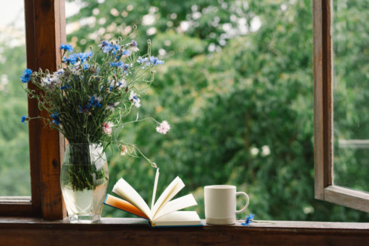 Open book, cup, and vase of flowers on a windowsill with greenery outside.